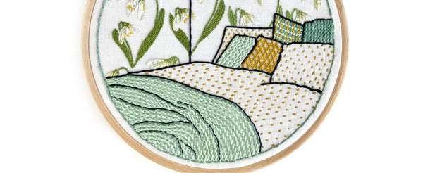 Embroidery hoop with a white background featuring a green and white floral pattern and a stitched bedroom scene with a bed, cushions, and a window.